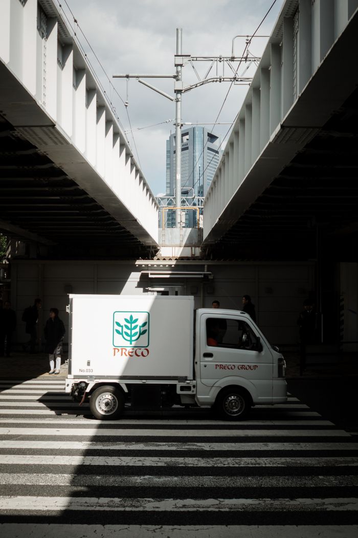 about-us-02 Mini truck passes under railway bridge in Tokyo's Shiba district with a modern skyscraper view.