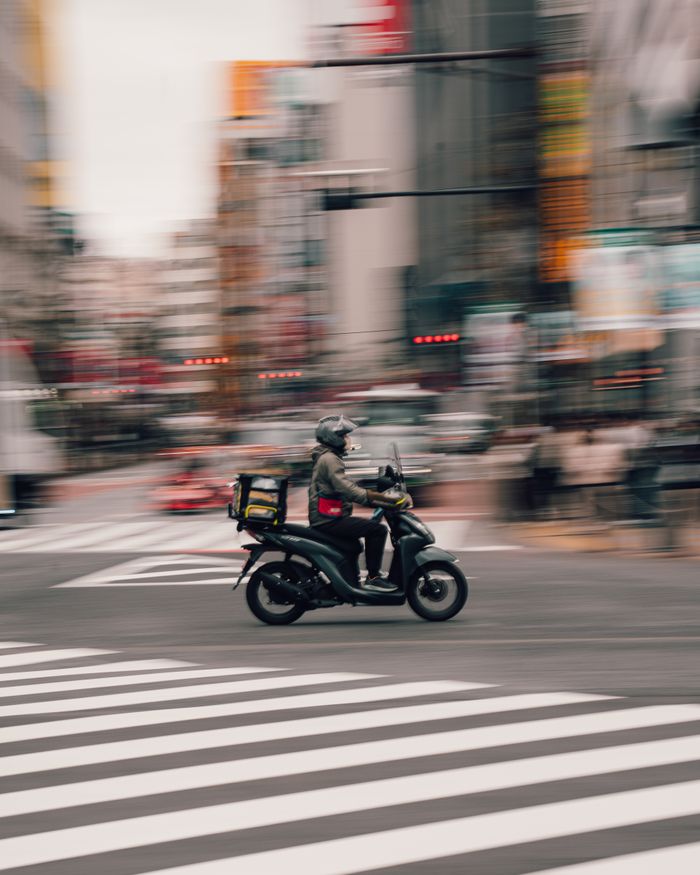 services-02 A delivery rider on a scooter speeds through a busy city street in Osaka, Japan, capturing urban hustle and motion.