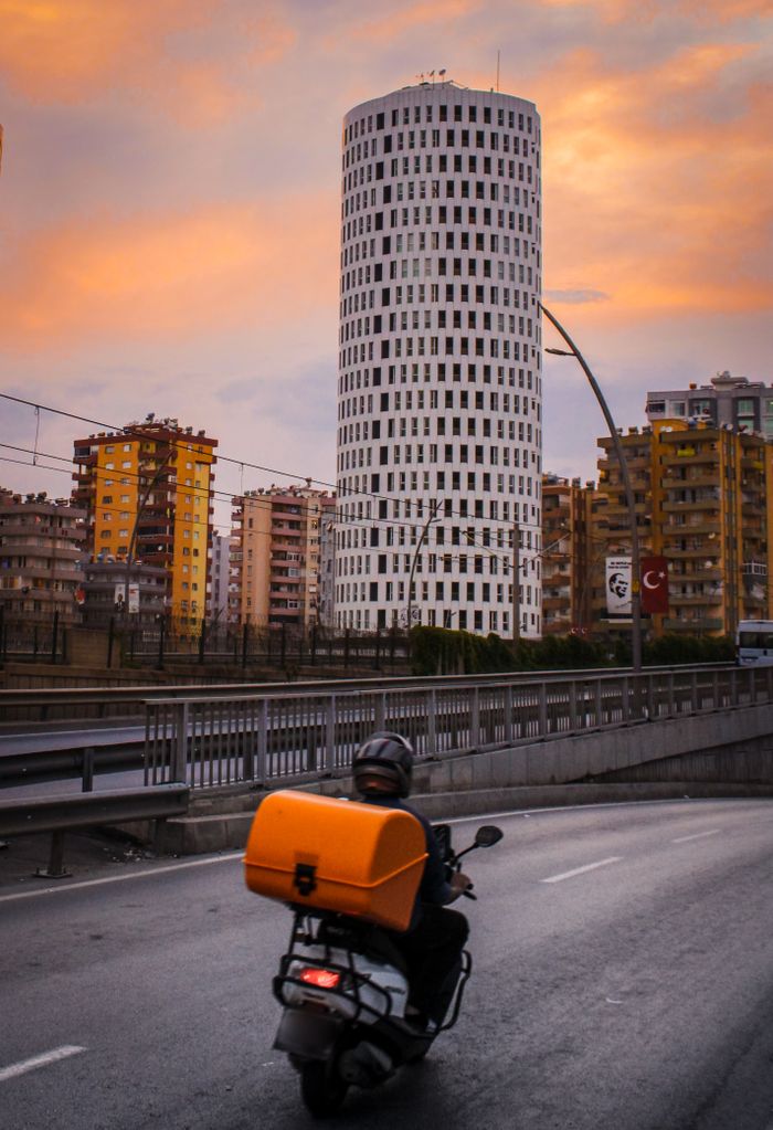 Motor scooter rider on a city road with a high-rise building at sunset.